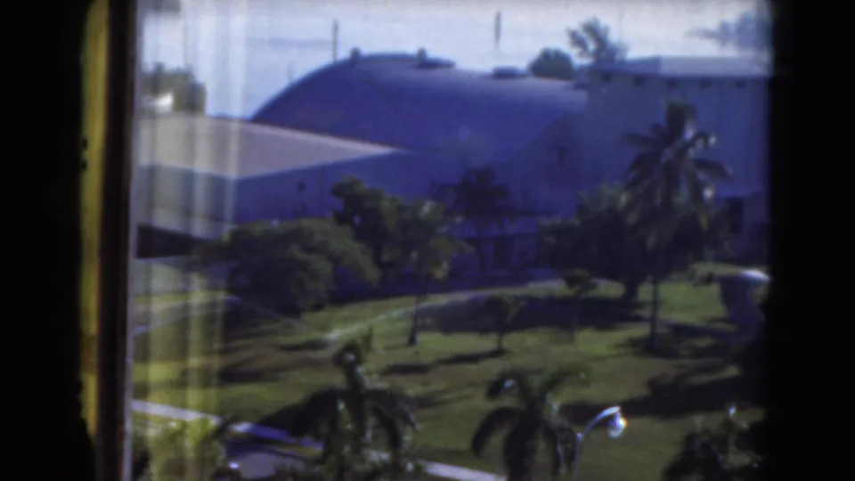 1975 Florida 8mm frame showing a sunlit tropical garden with palms and a large domed building beyond the window.