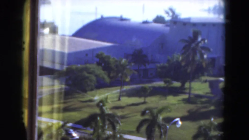 1975 Florida home-movie frame looking through a window at palms, lawn, and a large pale building with a domed roof.