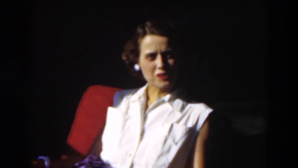 1975 home-movie frame of an adult woman in a white sleeveless blouse seated in a red chair, looking toward the camera.