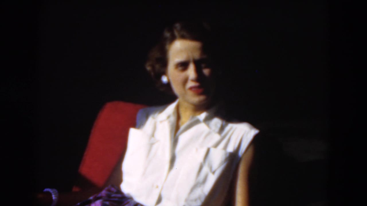 Adult woman in a white sleeveless blouse sits in a red chair against a dark background.
