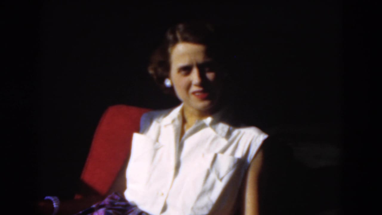 Adult woman in a white blouse sits in a red chair, lit from one side.