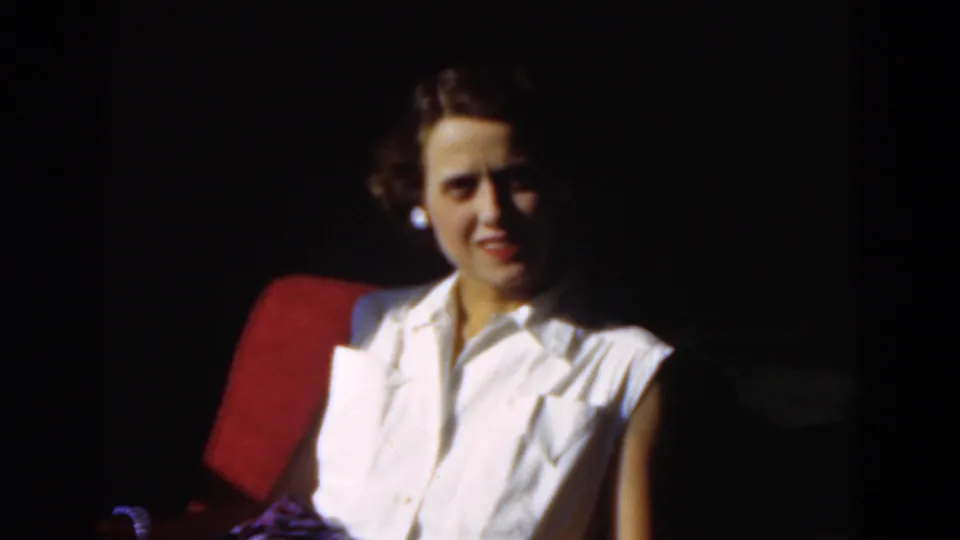 A seated adult woman in a white blouse begins to smile toward the camera in this 1975 Florida 8mm frame.