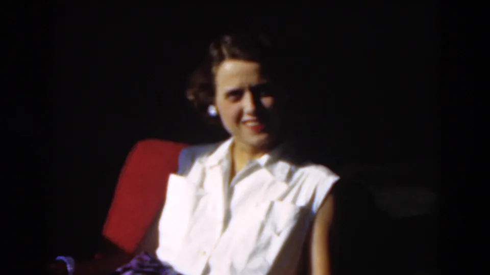 The 1975 Florida home-movie frame shows the woman smiling warmly from the red chair, with her white blouse catching the light.
