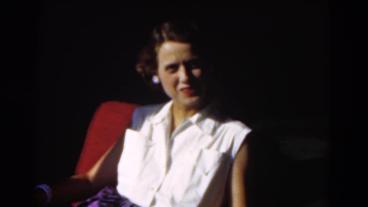 Young woman in a white sleeveless blouse smiles from a red chair in 1975 Florida home-movie footage.