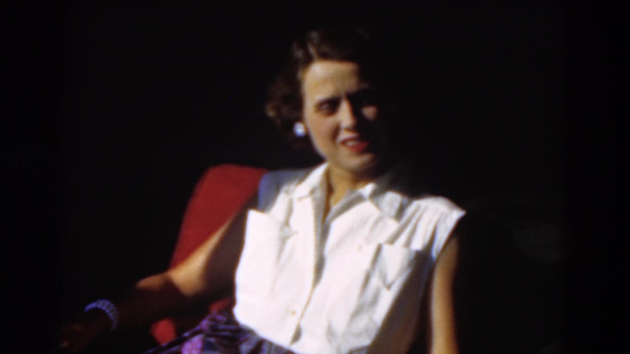 Young woman in 1970s clothing sits in a red chair, softly lit against a dark background.