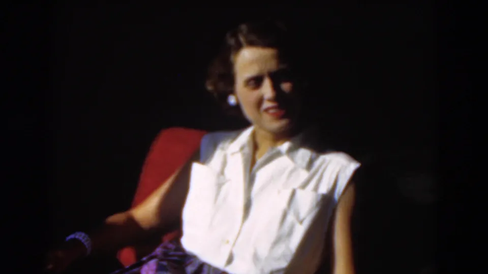 Young woman in 1970s clothing sits in a red chair, softly lit against a dark background.