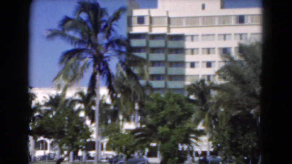 Florida 8mm frame of a white multi-story building behind palms, with greenery and small cars visible near the base.