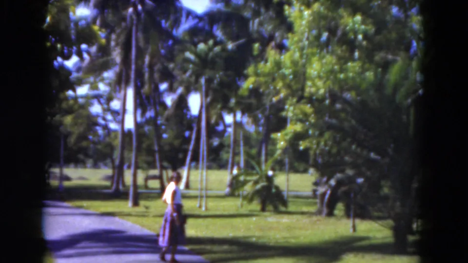 1975 Florida 8mm frame showing a woman near a paved path amid tall palms, lawn, and dense tropical greenery.