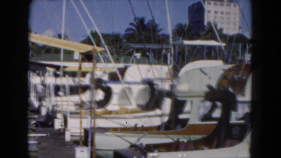 1975 Florida 8mm marina frame showing docked boats, masts, a distant building, and palm trees beyond the water.