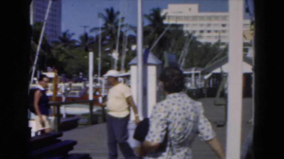 1975 Florida 8mm frame showing casually dressed adults walking on a marina dock near boats, masts, and palm trees.