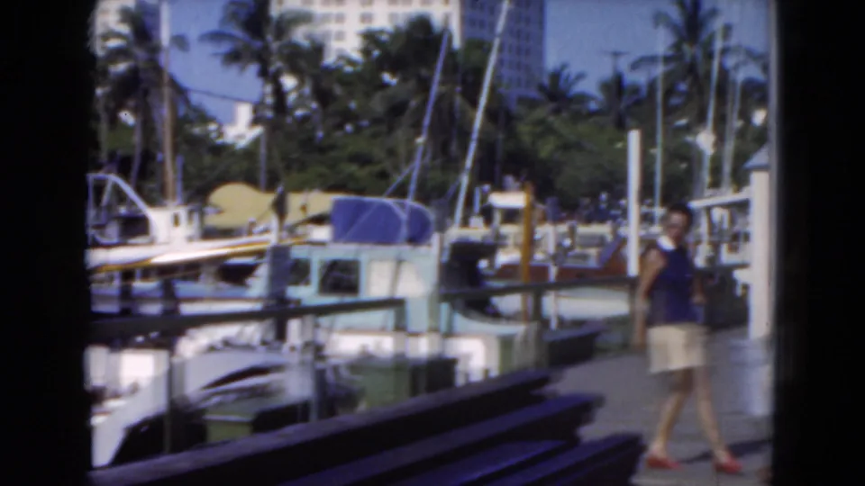 1975 Florida marina frame with a man near benches and docked boats, with masts, palm trees, and waterfront structures visible.