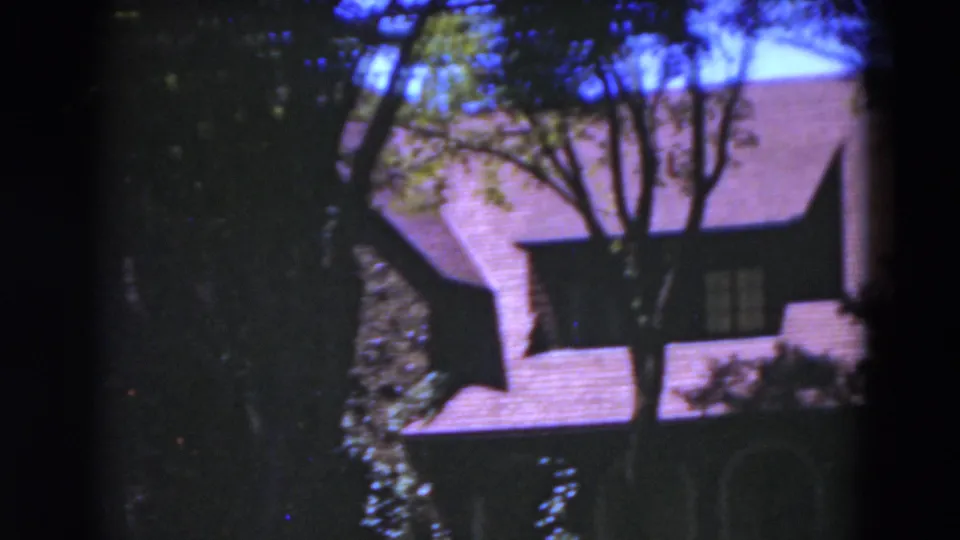 1975 Florida 8mm frame with a Tudor-style roof, window, tree trunks, and shaded wall details of the residence.