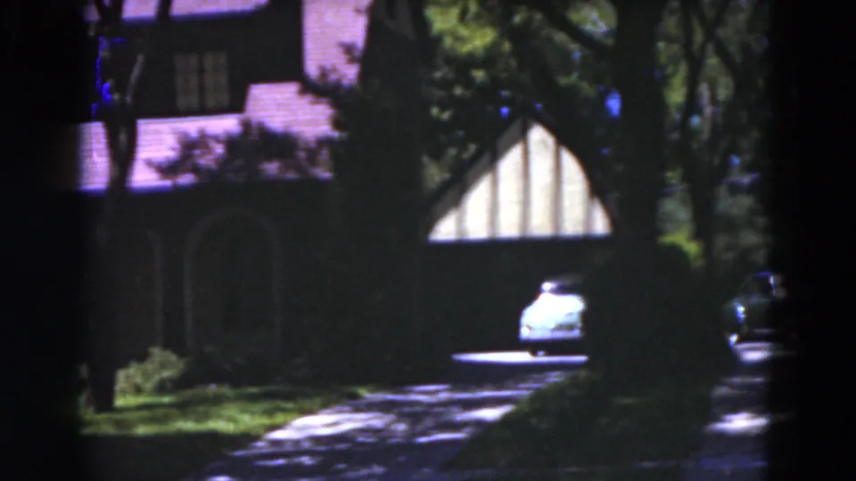 1975 Florida home-movie frame of a shaded house exterior with a driveway, trees and a light-colored car near the right side.