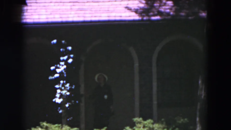1975 Florida home-movie frame with a woman standing in a dark arched doorway, partially framed by shrubs and the house facade.