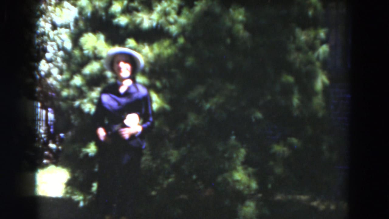 Woman in hat stands before dense green foliage while facing the camera.