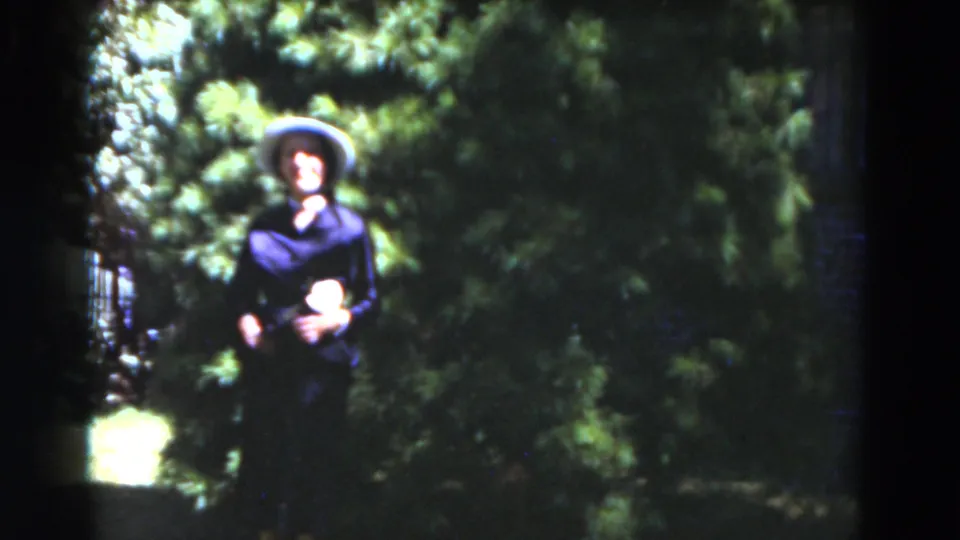 A woman in a wide-brimmed hat and dark dress faces the camera in a lush Florida garden, with bright greenery behind her.