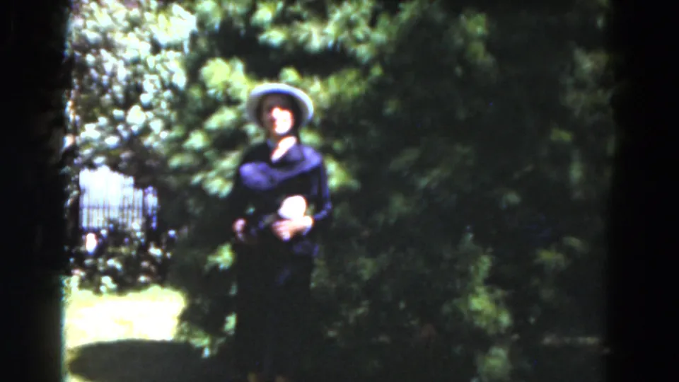 The woman remains posed in the garden, framed by a fence or railing, sunlit lawn, and dense greenery in 1975 Florida footage.