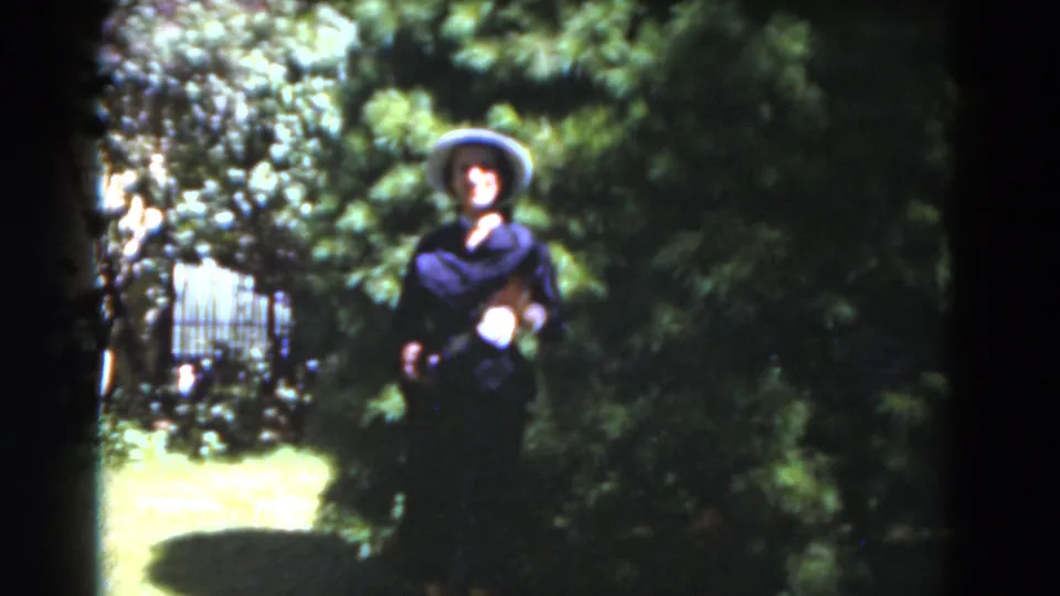 A woman in a broad hat appears to wave toward the camera in a Florida garden, surrounded by trees, lawn, and soft 8mm color.