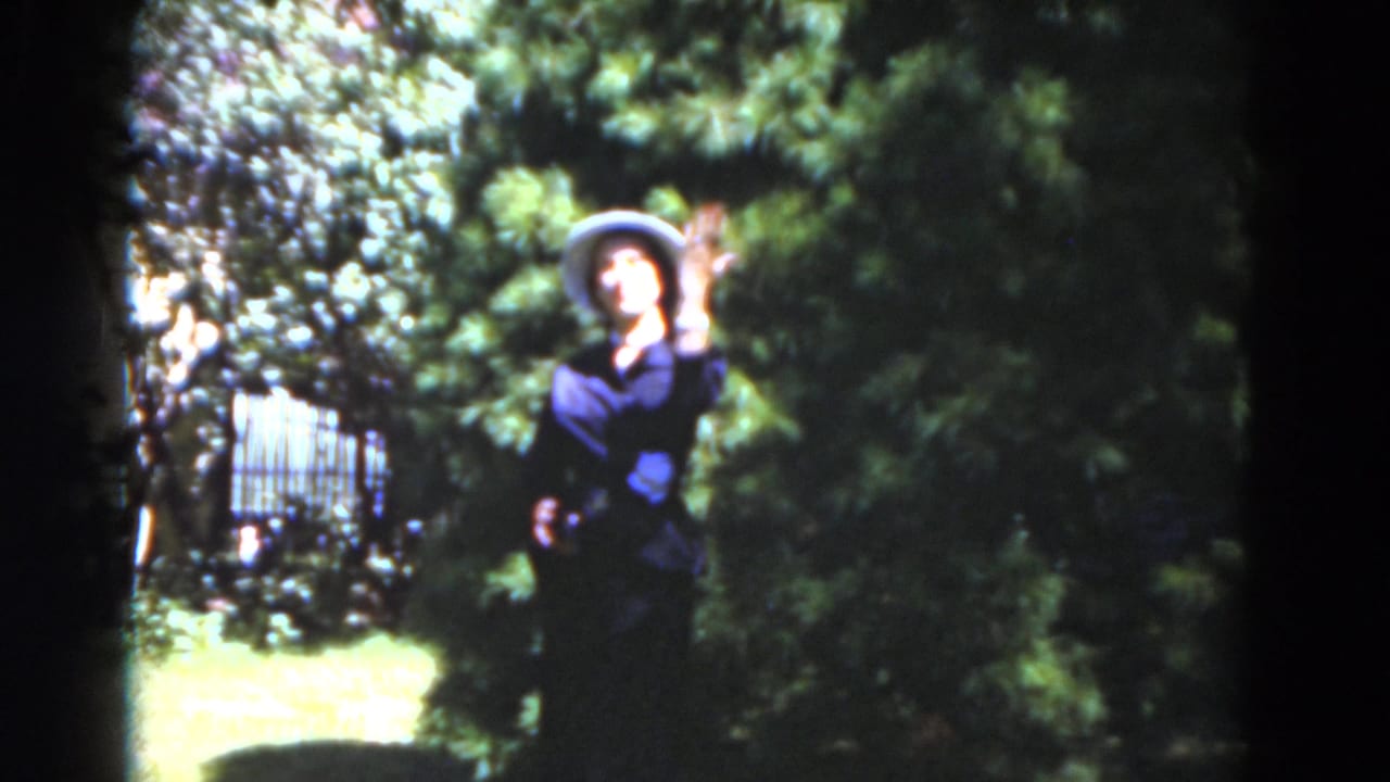Woman in a light hat waves while standing in a sunlit garden.