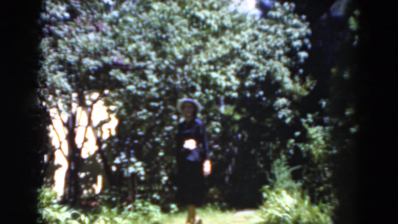 Woman in a dark suit and wide-brimmed hat walking on a garden path.