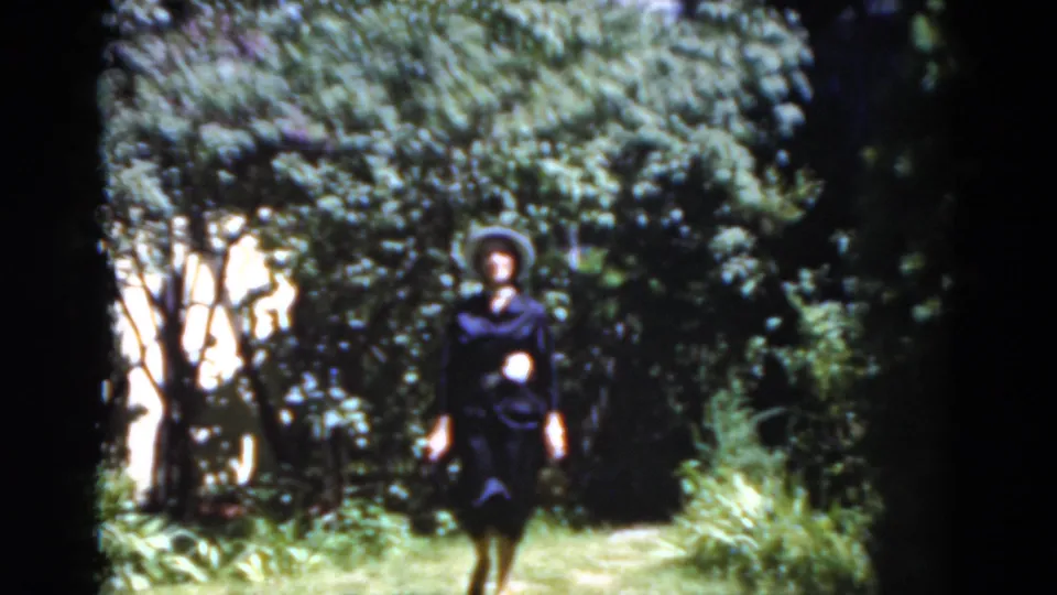 A woman in formal dark clothing walks closer along a Florida garden path in 1975 home-movie footage.