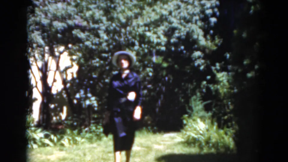The woman appears close to the camera on a leafy Florida garden path, wearing a dark suit and wide-brimmed hat.