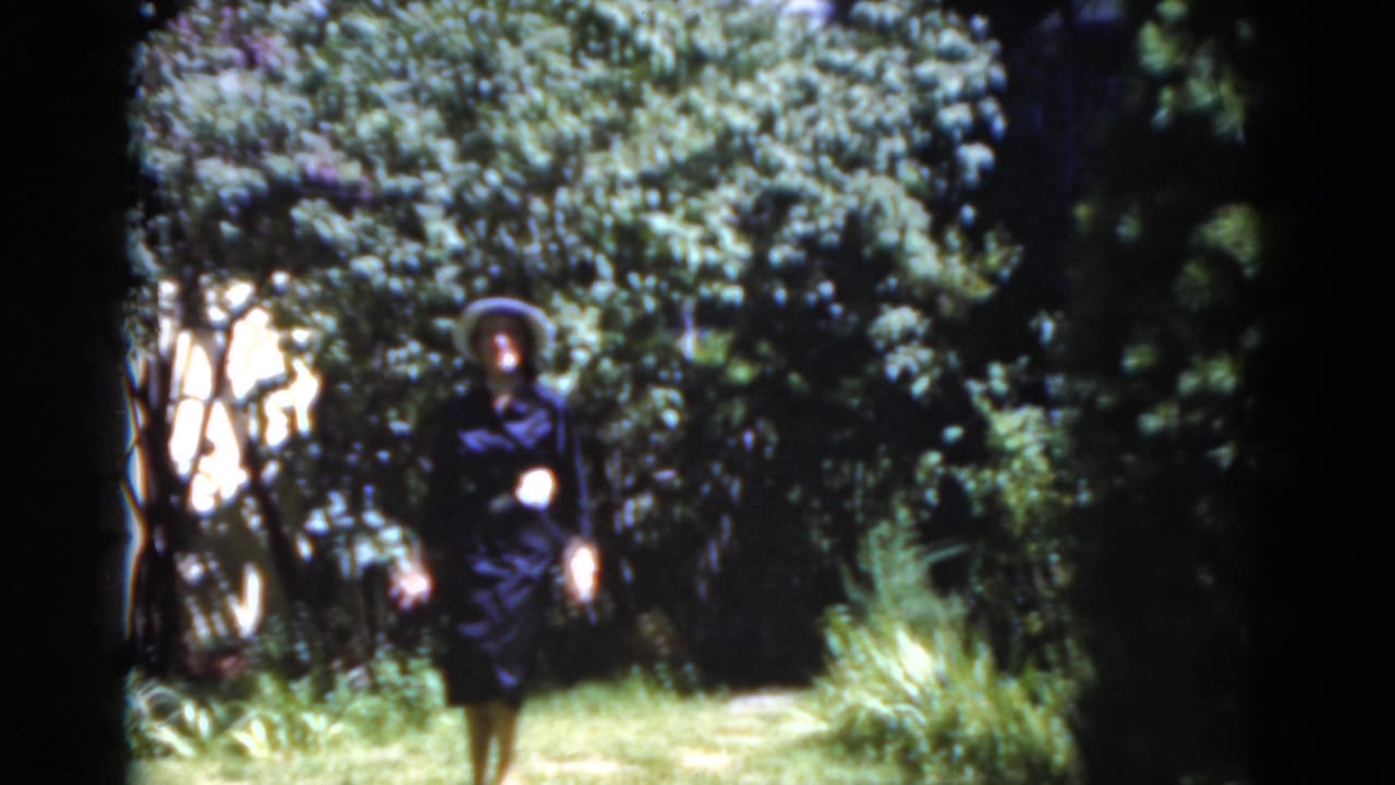 Woman in a dark dress and wide-brimmed hat walking along a garden path.