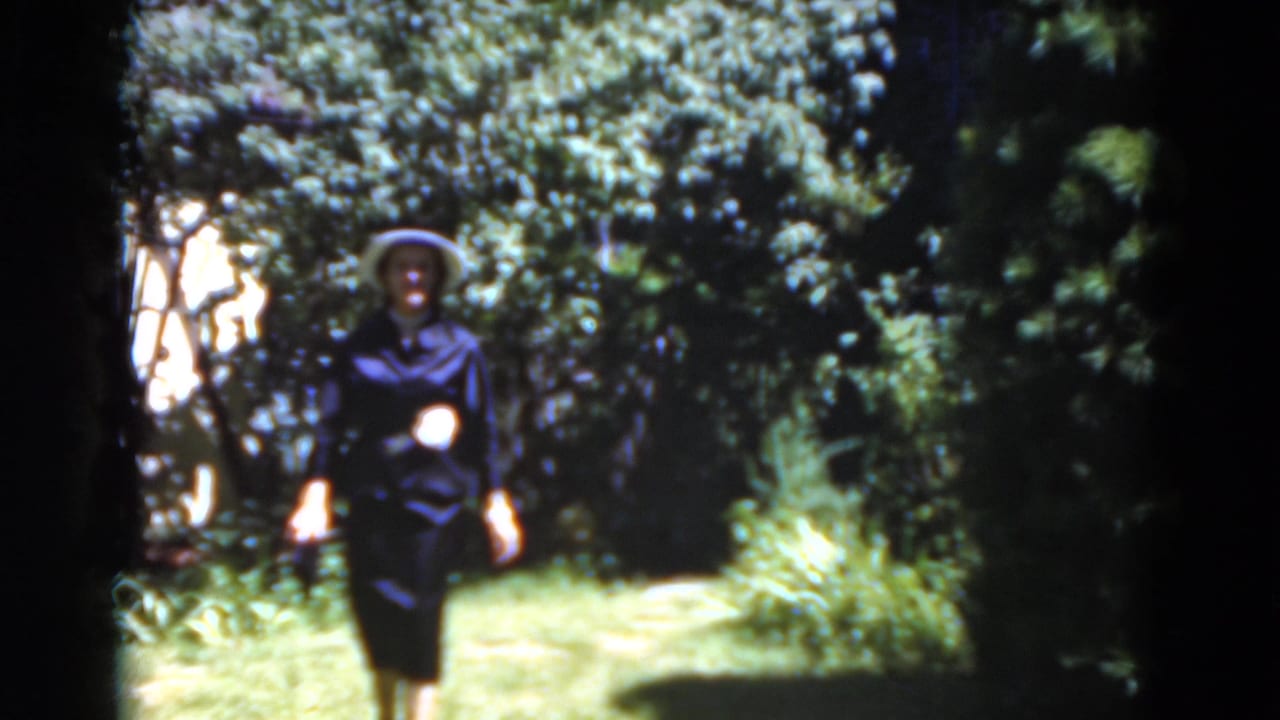 Woman in a hat walking closer on a bright garden path surrounded by trees.