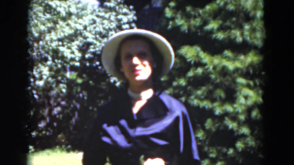 Close 1975 Florida home-movie view of the woman smiling in a light hat, framed by dark trees and bright garden foliage.