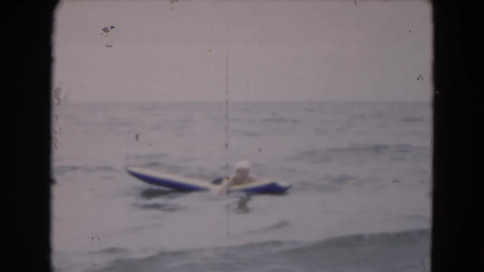 In 1975 Florida home-movie footage, a woman in a swim cap floats on a blue-and-white raft in calm ocean water.