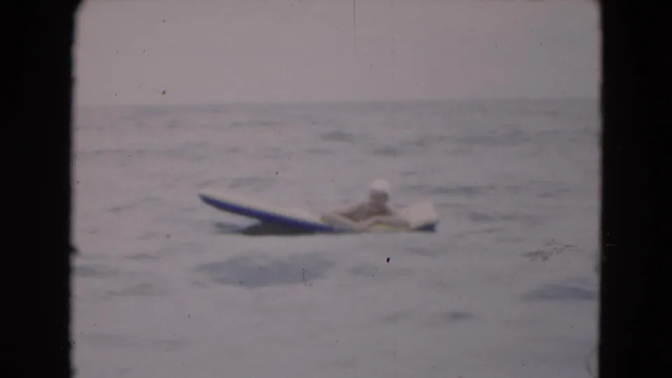 A woman in a swim cap lies low on a blue-and-white raft, with small waves and faded 8mm texture visible.
