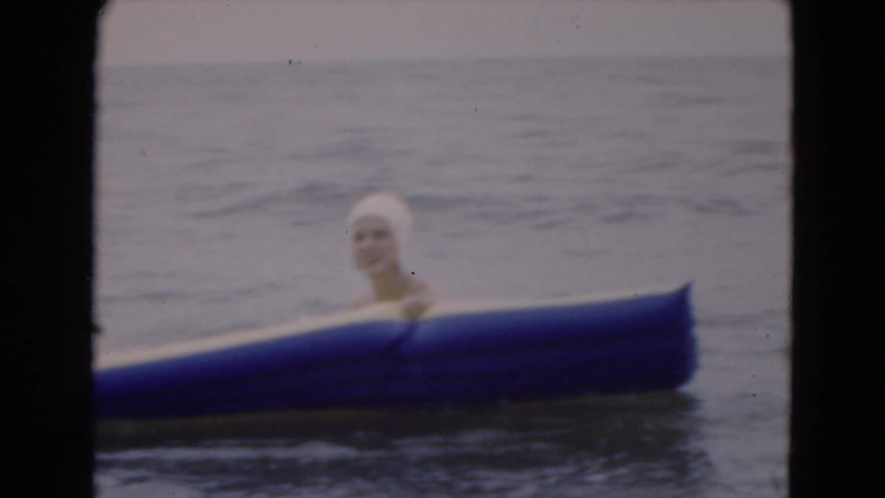1975 Florida 8mm frame of a woman in a white swim cap partly hidden behind a blue inflatable raft in the ocean.