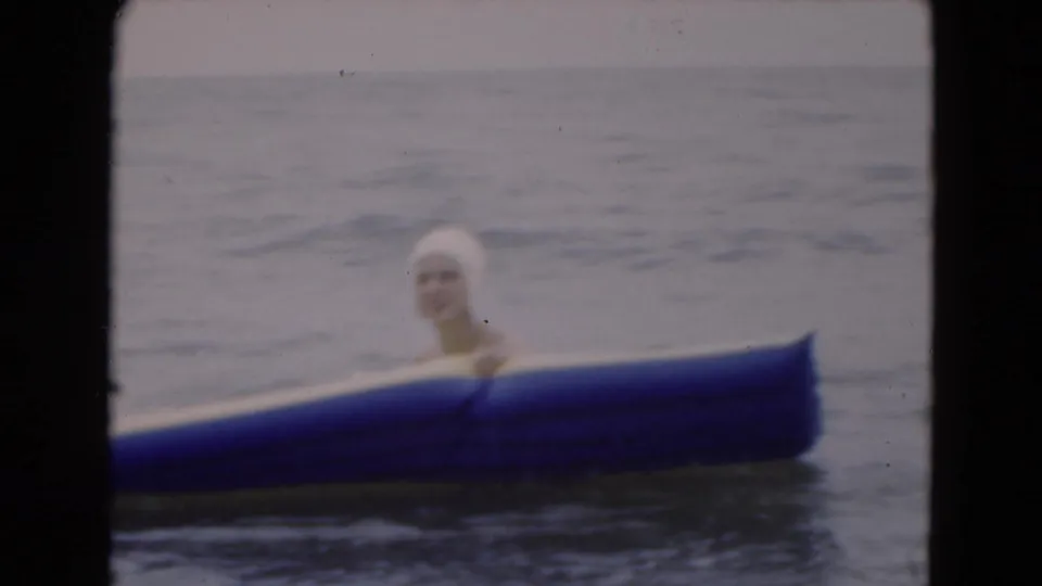 1975 Florida 8mm frame of a woman in a white swim cap partly hidden behind a blue inflatable raft in the ocean.