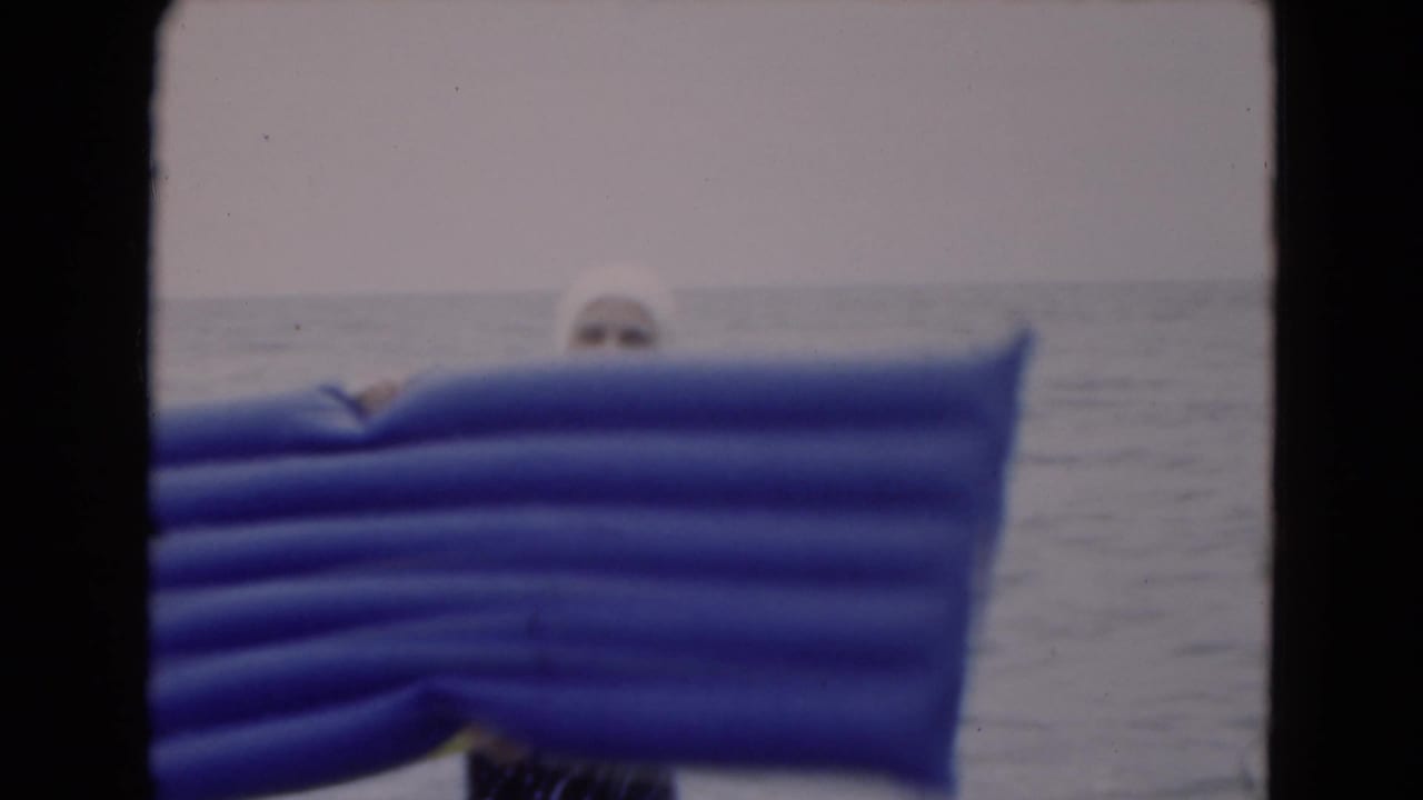 1975 Florida beach footage frame with a woman mostly obscured by a large blue inflatable raft in ocean water.