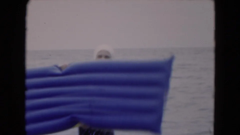 1975 Florida beach footage frame with a woman mostly obscured by a large blue inflatable raft in ocean water.