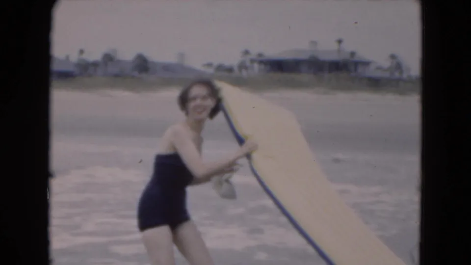 1975 Florida 8mm frame of a smiling woman in a dark one-piece swimsuit holding a yellow inflatable raft near the beach.