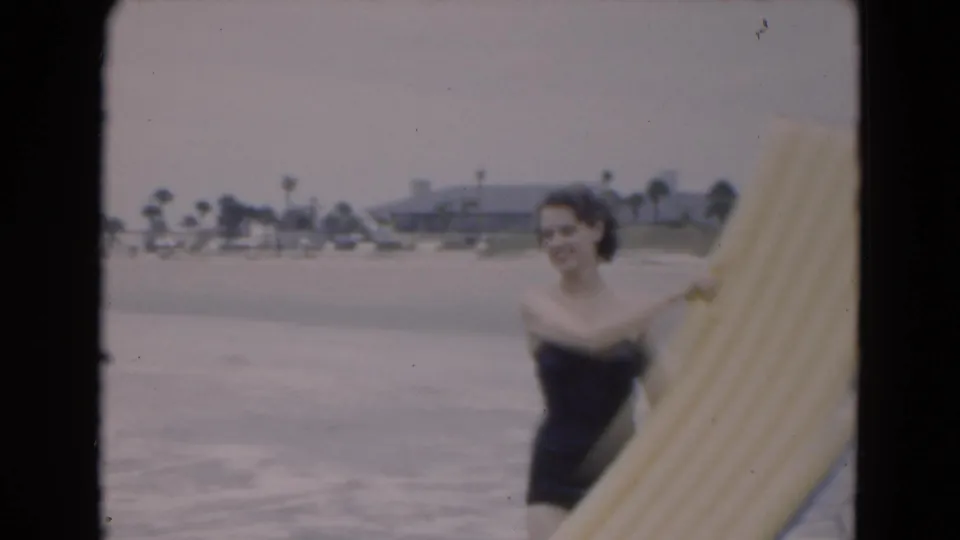 1975 Florida 8mm frame of a woman in a dark swimsuit carrying a yellow inflatable raft with surf, palm trees, and buildings behind her.