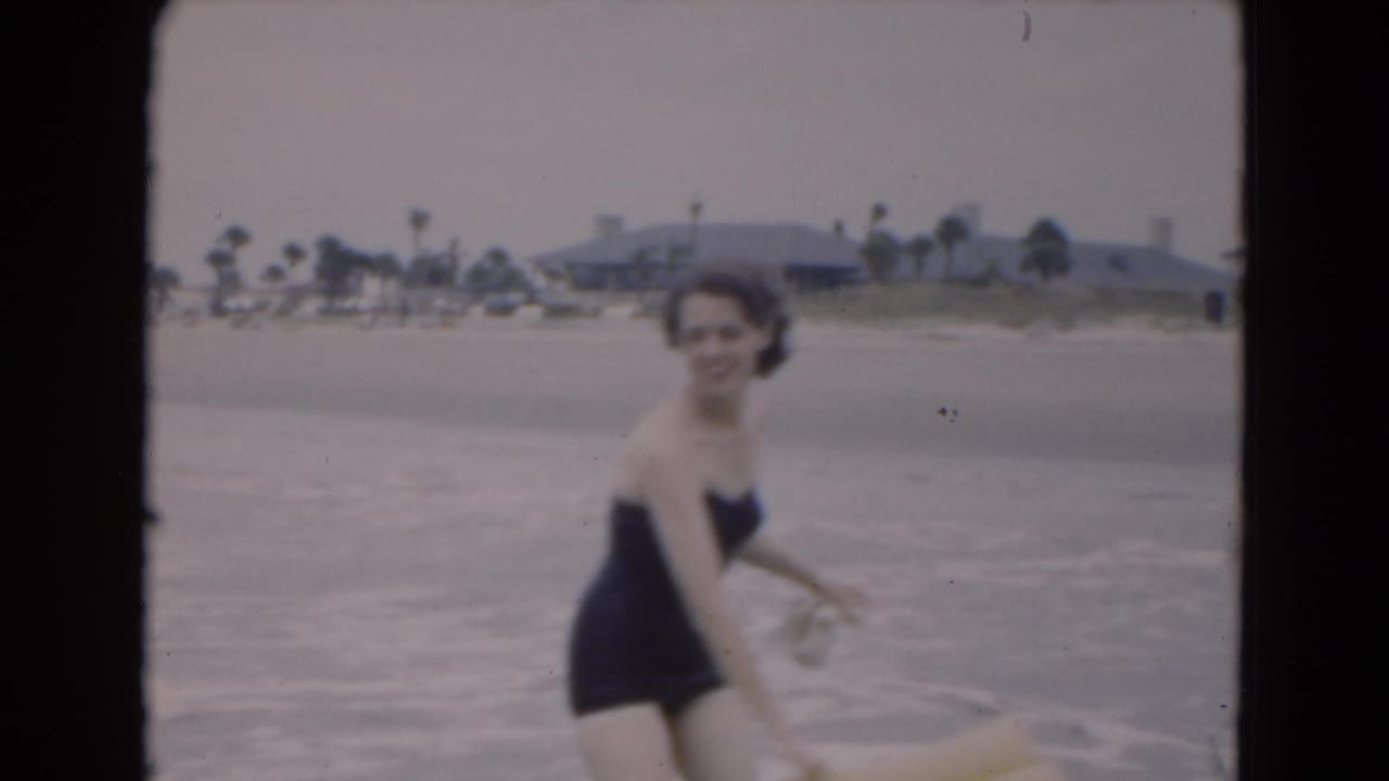 Woman in a dark one-piece swimsuit stands in shallow surf holding a yellow inflatable raft.