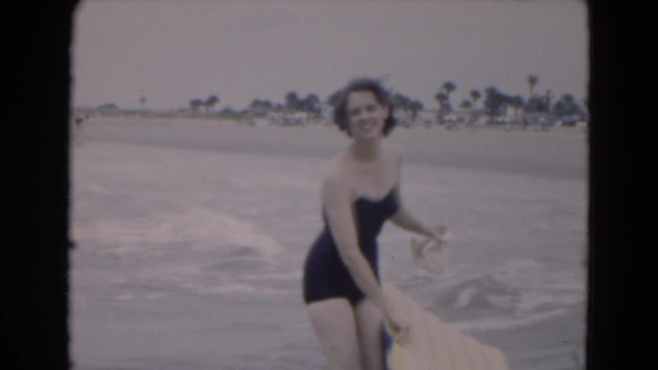 Smiling woman in shallow waves holds an inflatable raft with beach and palms behind her.