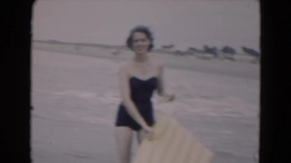 The woman faces the home-movie camera in shallow beach water, holding a yellow inflatable raft as the shoreline recedes behind her.