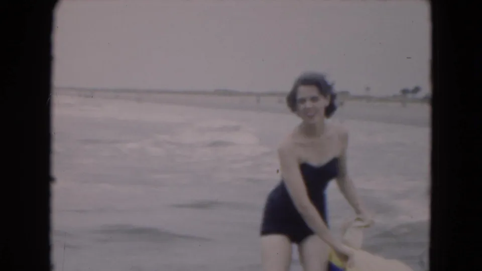A woman in a dark swimsuit bends toward a yellow raft in the shallow ocean, with soft waves and the sandy Florida shore behind her.