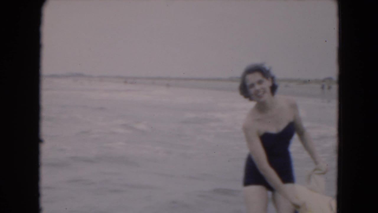 In 1975 Florida home-movie footage, a woman in a dark one-piece swimsuit smiles near the waterline on an overcast beach.