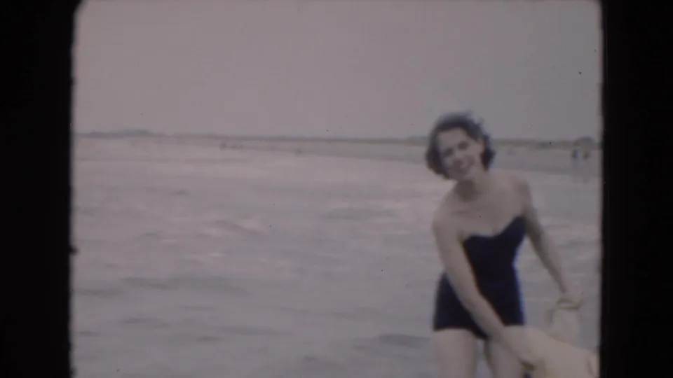 In 1975 Florida home-movie footage, a woman in a dark one-piece swimsuit smiles near the waterline on an overcast beach.