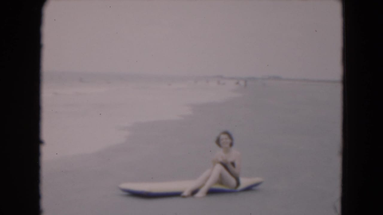 The woman sits sideways on a surfboard at the Florida shoreline, with gentle waves and distant beachgoers behind her.