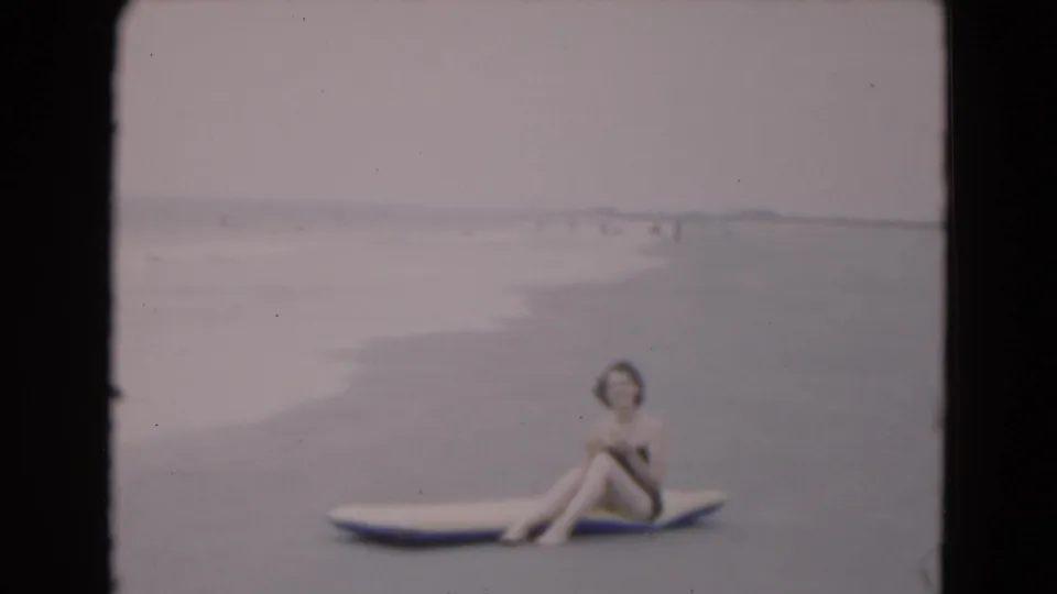 The woman sits sideways on a surfboard at the Florida shoreline, with gentle waves and distant beachgoers behind her.