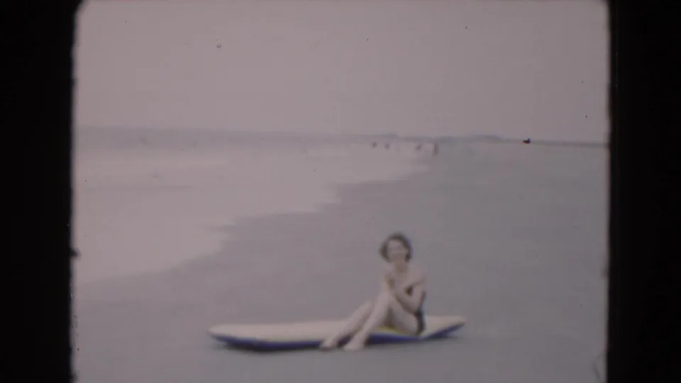 A seated woman poses on a surfboard in shallow Florida surf, with pale ocean water and small distant figures in the background.