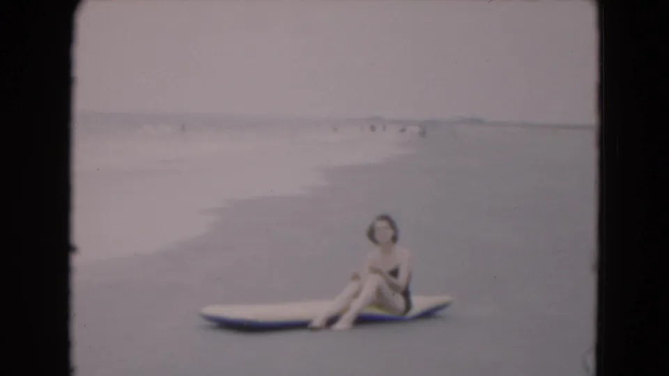 The woman remains seated on the surfboard at the Florida beach, framed by shallow surf, muted sky, and far-off beach visitors.