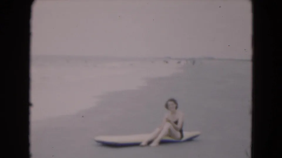 In 1975 Florida beach footage, a woman in a dark one-piece swimsuit sits on a yellow-and-blue raft at the edge of the ocean.