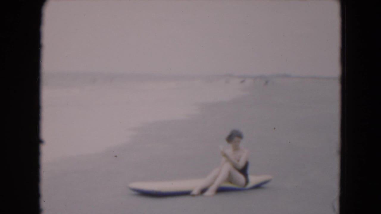A woman in a dark swimsuit sits on an inflatable raft along the Florida shoreline, turned slightly downward in soft 8mm focus.