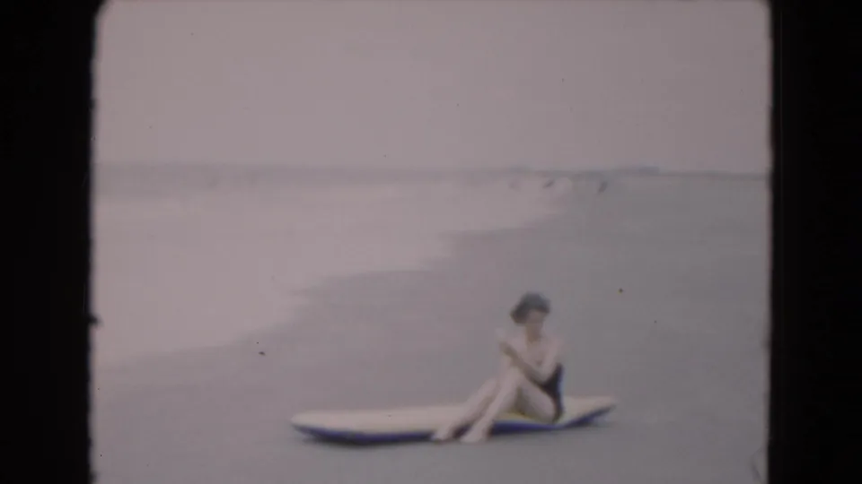 A woman in a dark swimsuit sits on an inflatable raft along the Florida shoreline, turned slightly downward in soft 8mm focus.